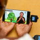 Two hands of the same person hold a phone screen showing a video lesson. The phone has a USB-like memory device attached to it.